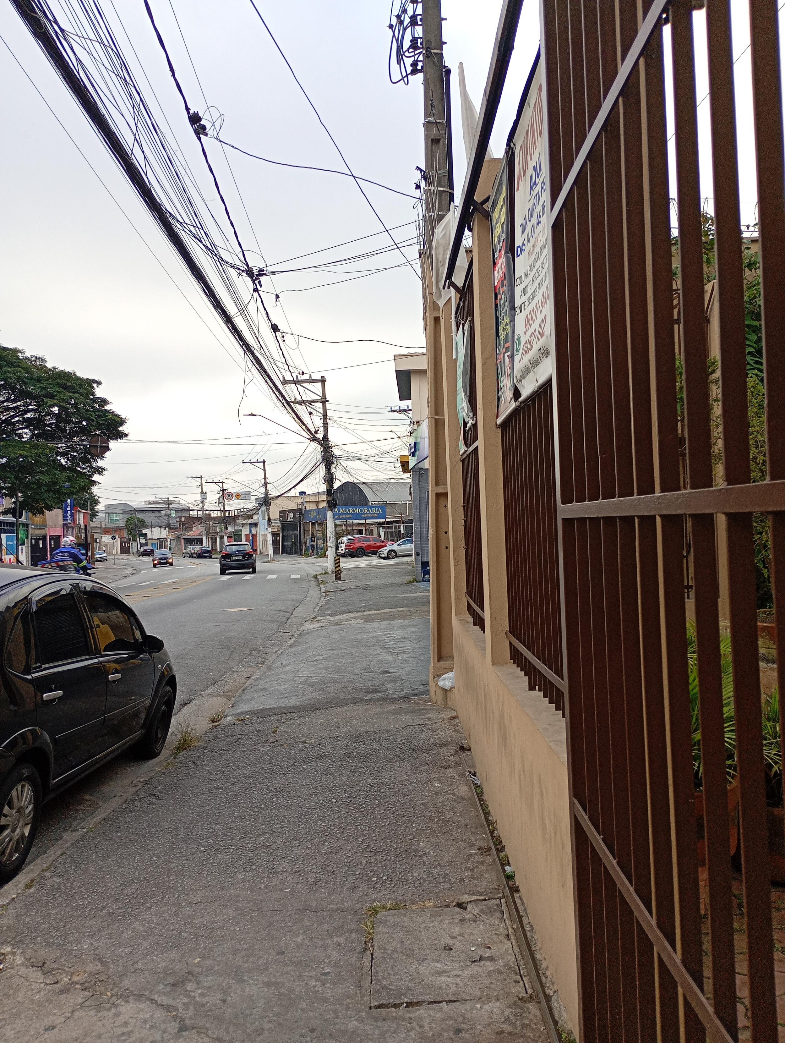 Uma foto em plano médio e ângulo normal mostra uma calçada de concreto cinza que se estende em direção ao fundo. À direita, há um portão e uma grade alta de metal marrom instalados sobre um muro bege claro, onde placas verticais com anúncios estão fixadas. À esquerda, parte de um carro preto está estacionada rente ao meio-fio, paralela à rua onde outros veículos transitam ao longe. No topo, uma rede densa de cabos e fios elétricos cruza o céu nublado, conectada a postes de energia ao longo da via urbana.