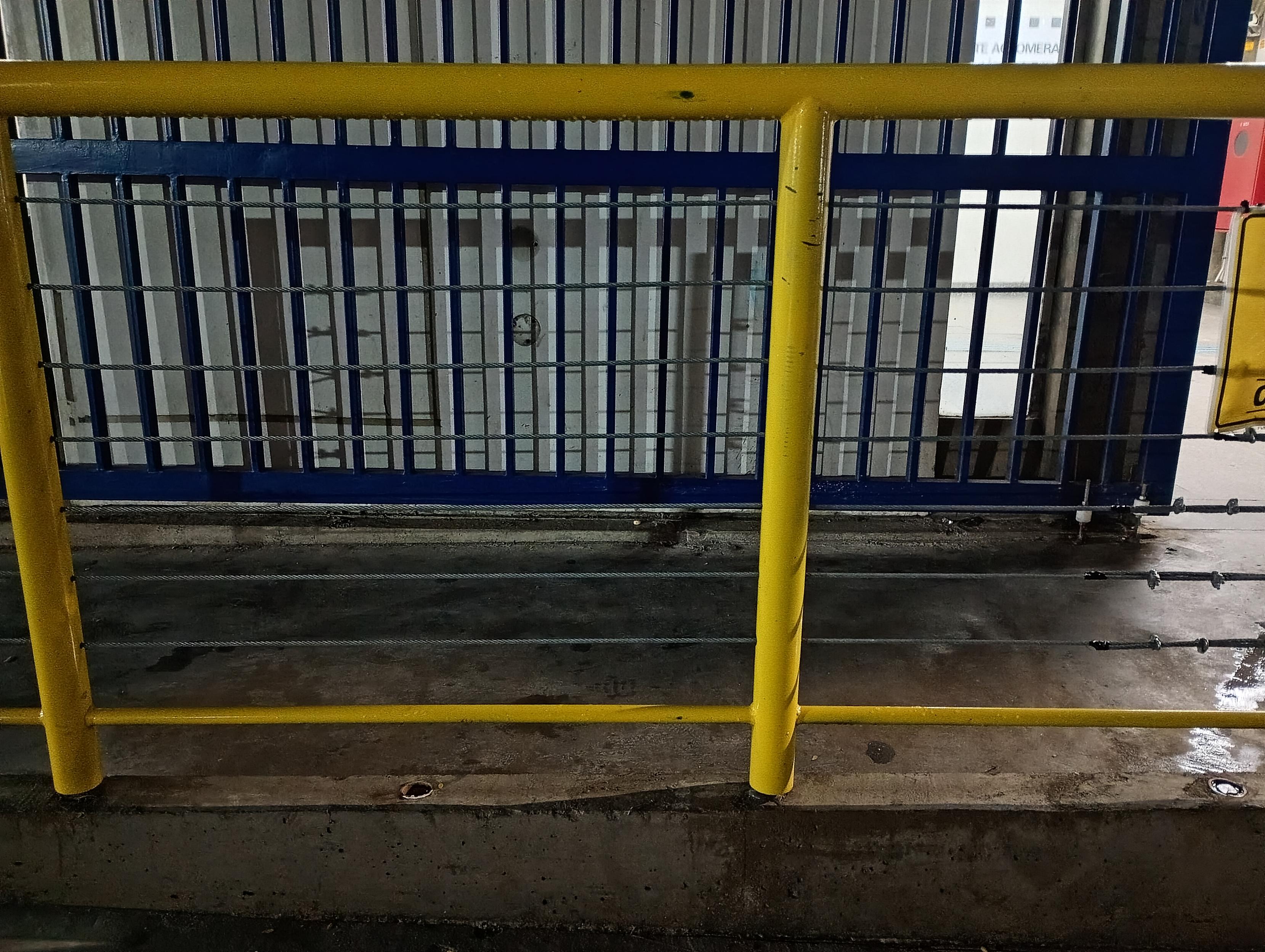 Foto noturna de uma estrutura metálica pintada de amarelo vibrante serve como guarda-corpo em primeiro plano. Ela é composta por tubos cilíndricos, incluindo um corrimão superior horizontal e colunas verticais que se fixam a uma mureta de concreto escuro e úmido no chão. Entre esses tubos amarelos, estendem-se horizontalmente finos cabos de aço paralelos. Logo atrás do guarda-corpo amarelo, há um portão ou grade de metal azul-escuro com barras verticais. Atrás das frestas da estrutura azul, avista-se uma parede encardida clara onde se projetam as sobras do guarda-corpo e das barras do portão, deixando tudo meio quadriculado.
